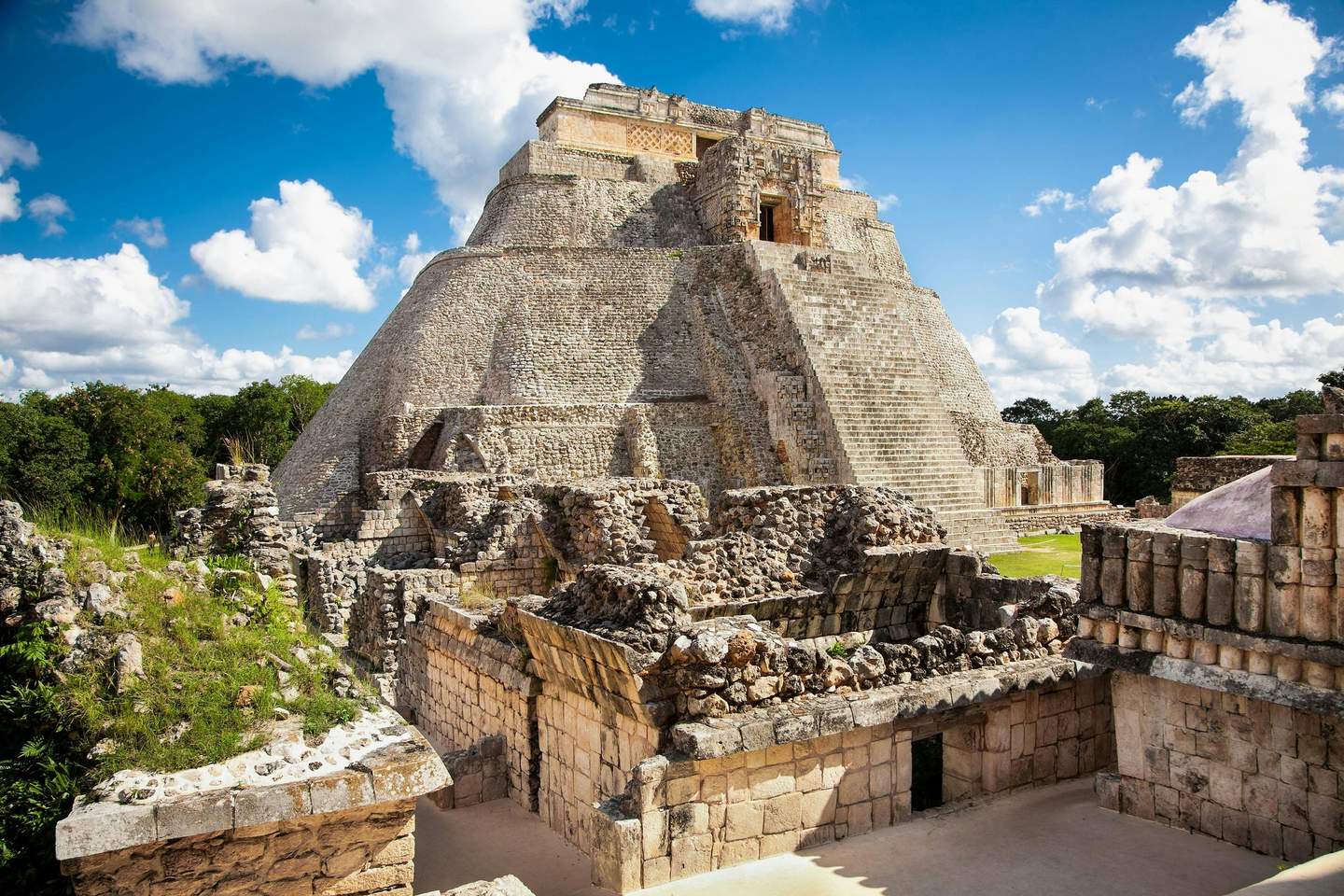 Uxmal Ruins Stone pyramid surrounded by ancient ruins under a blue sky with scattered clouds and lush green vegetation in the background.