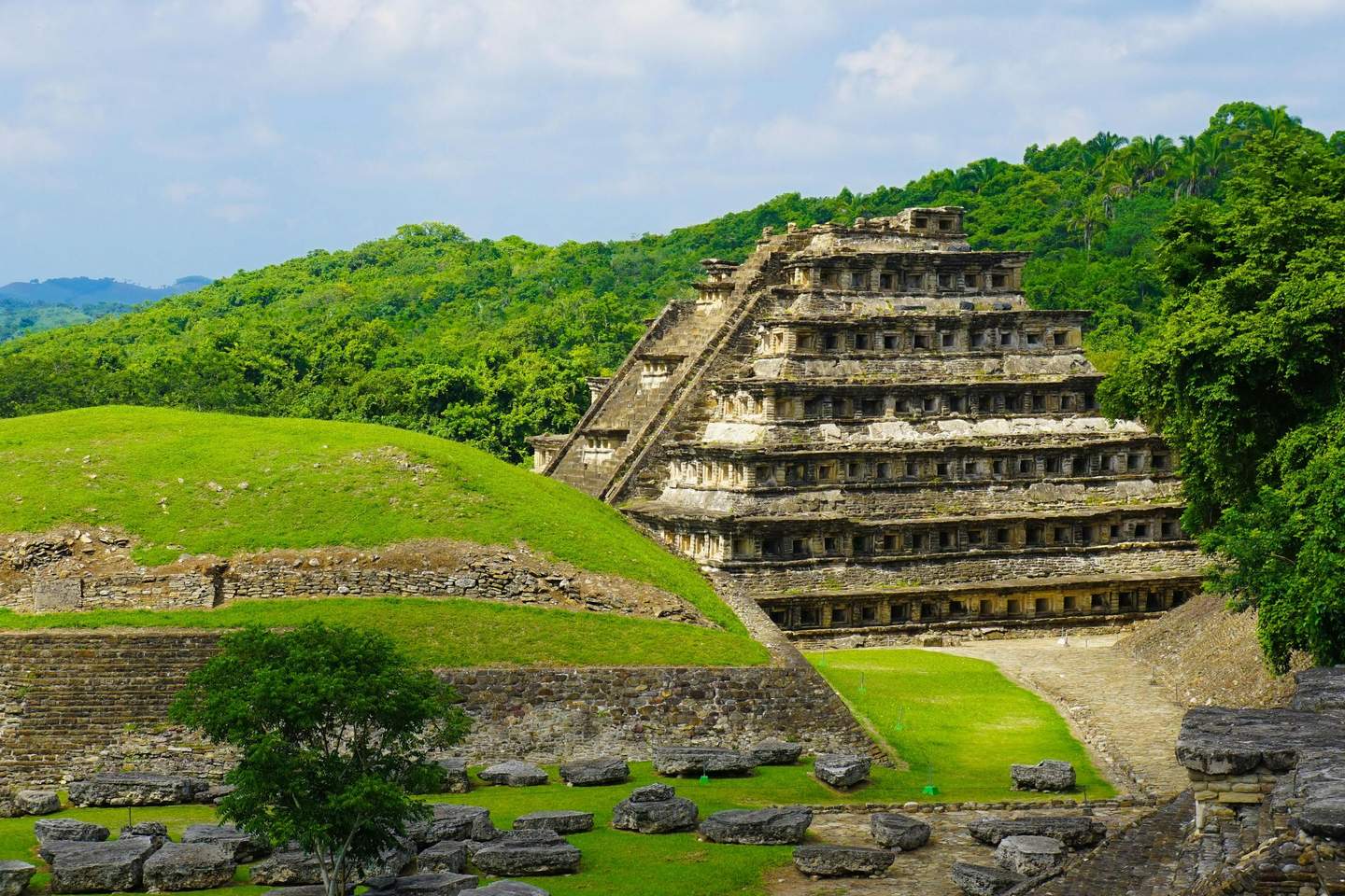 El Tajín A multi-tiered ancient pyramid stands in a green landscape with surrounding ruins and dense forest in the background.