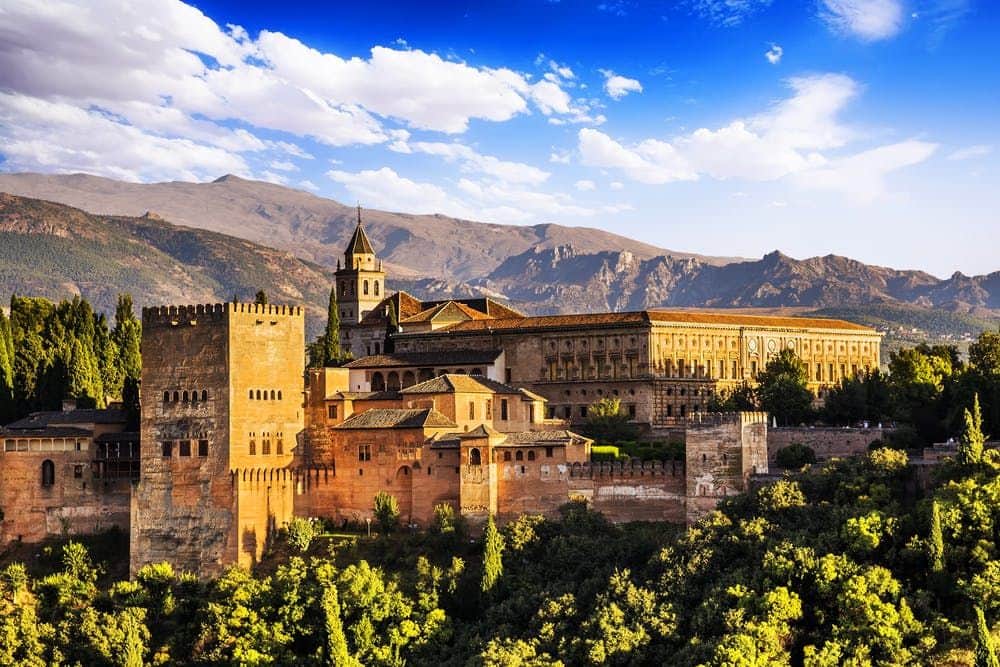 Alhambra A large, historic fortress with towers and ornate buildings sits amid lush greenery, with mountains and a blue sky in the background.