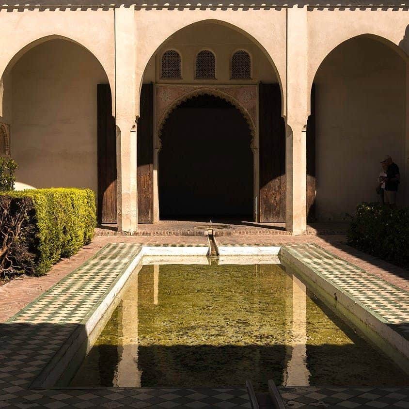 Alcazaba of Málaga A courtyard with an arched entrance, shallow pool, and greenery; a person stands in the shaded corner.