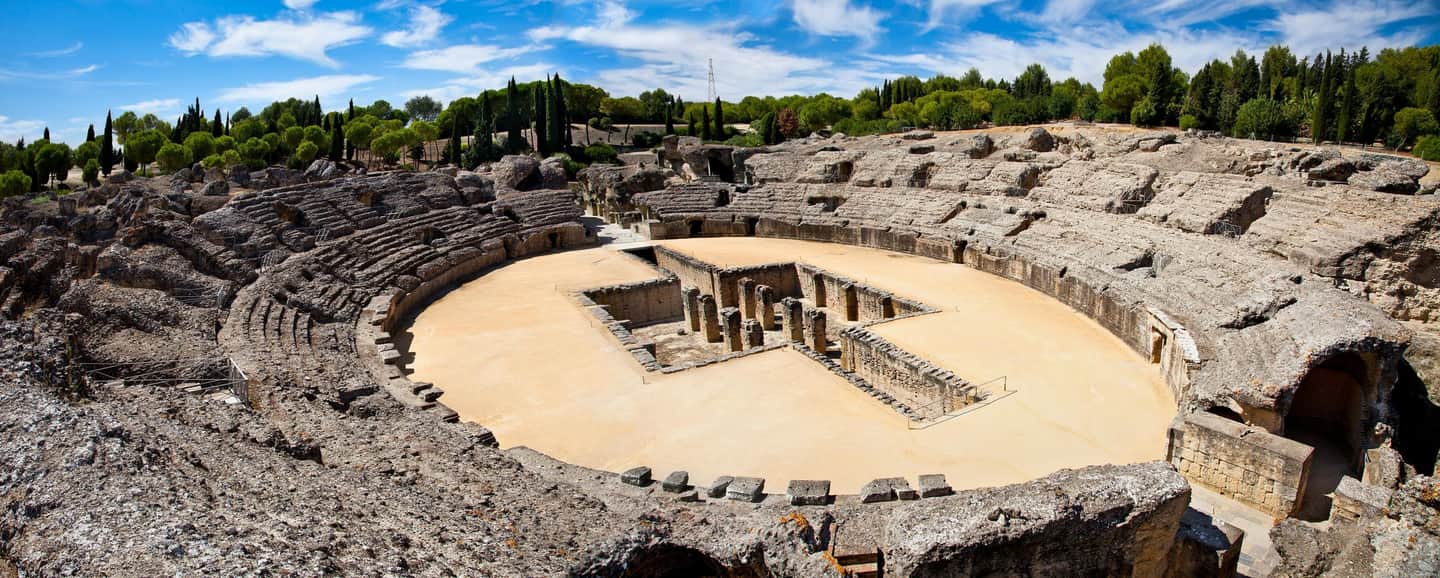 Itálica An ancient amphitheater with tiered seating, a central arena, and surrounding greenery under a partly cloudy sky.