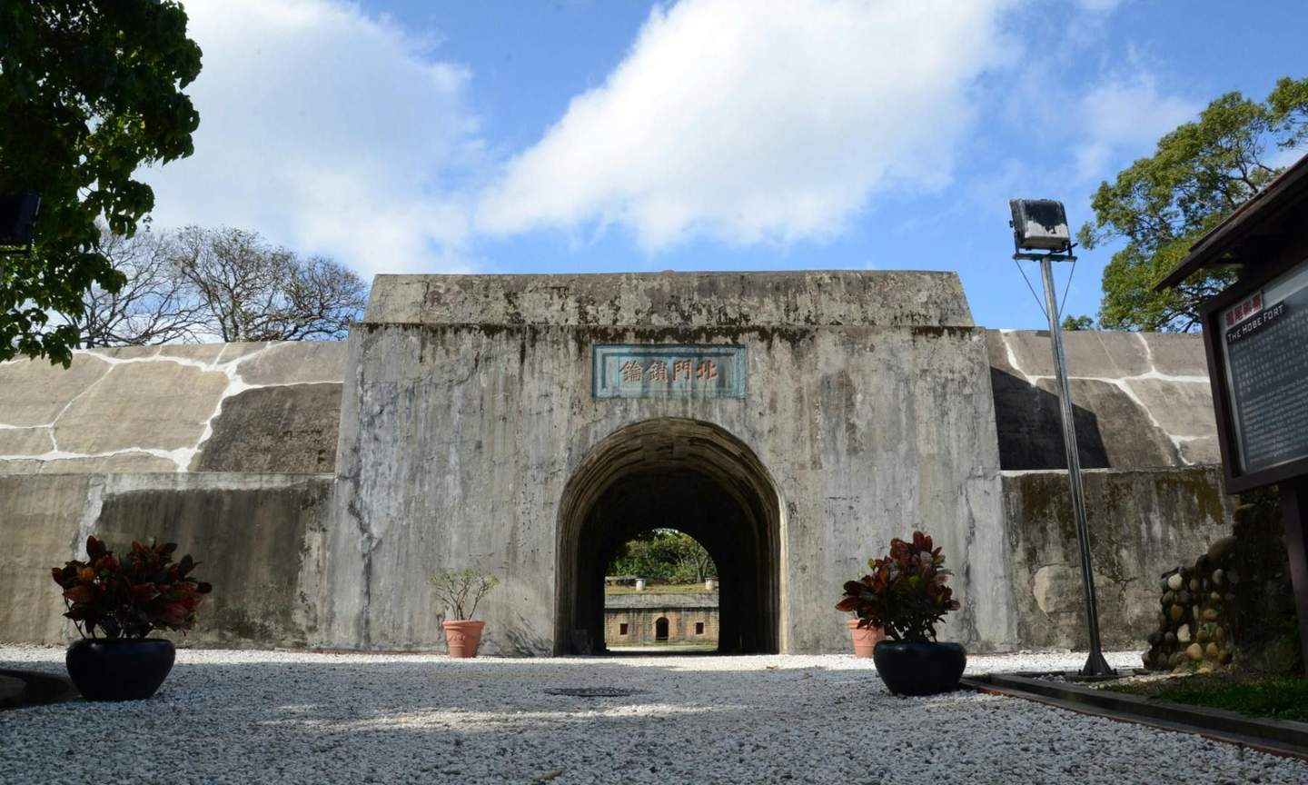 A concrete gate with an archway, surrounded by potted plants, leading to an inner courtyard under a blue sky.