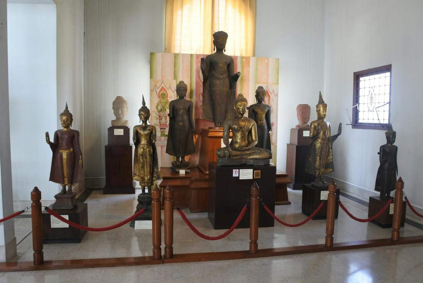 Room with various Buddhist statues on display, including standing, seated, and head sculptures, enclosed by red rope barriers.