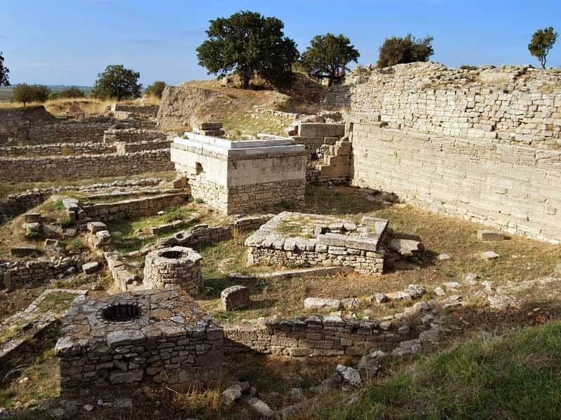 Trojan Ruins Ancient stone ruins with fragmented walls and structures, surrounded by scattered stones and sparse trees under a clear sky.