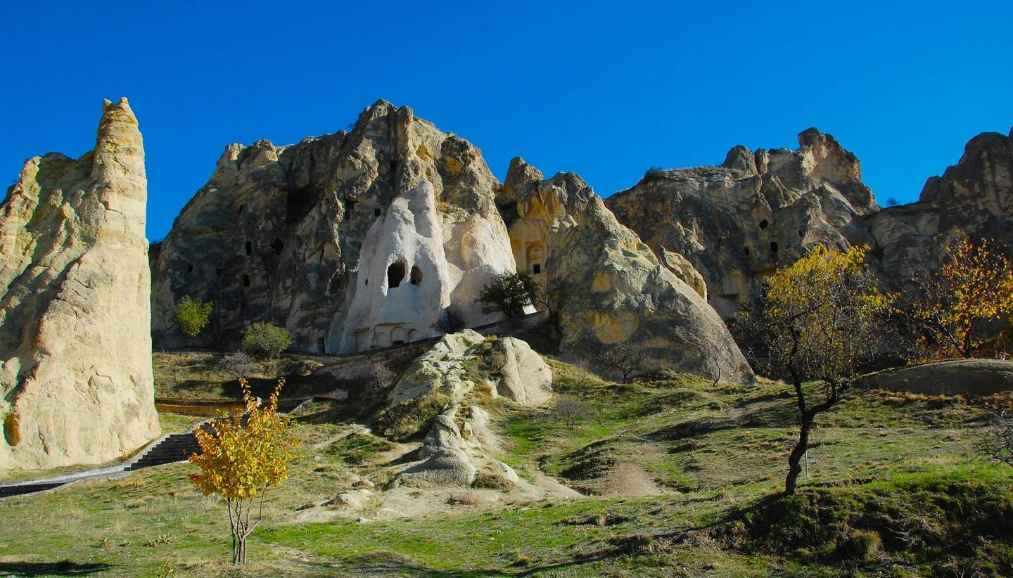 Göreme Open-Air Museum Rocky landscape with caves, sparse vegetation, and a clear blue sky. A path and some small trees are visible in the foreground.