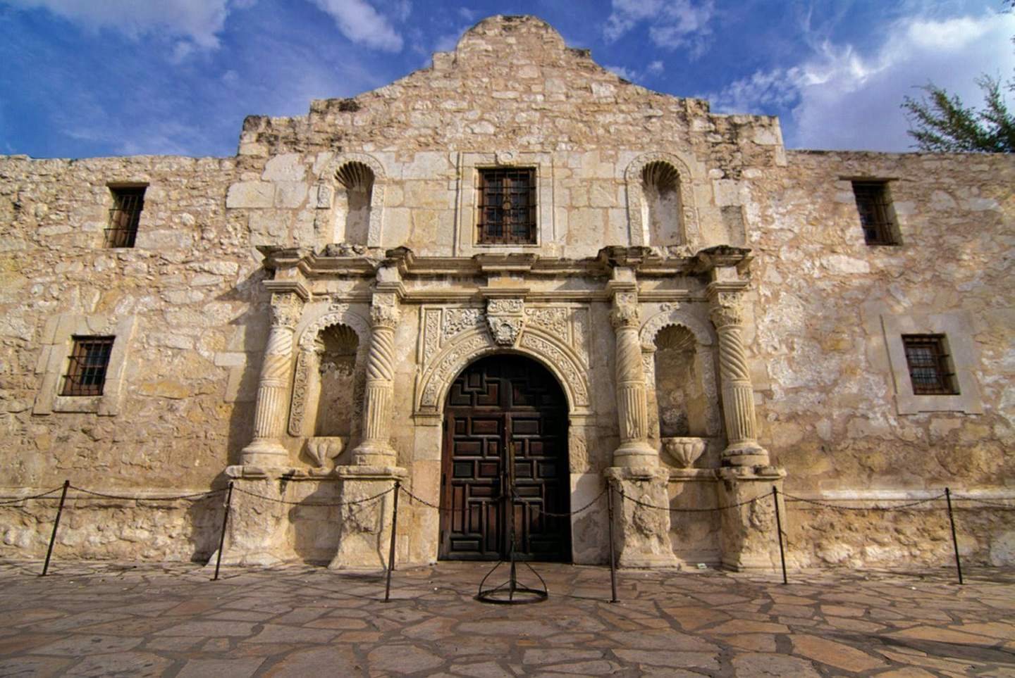 The Alamo Historic stone building facade with arched wooden door, columns, decorative carvings, barred windows, and partly cloudy sky above.