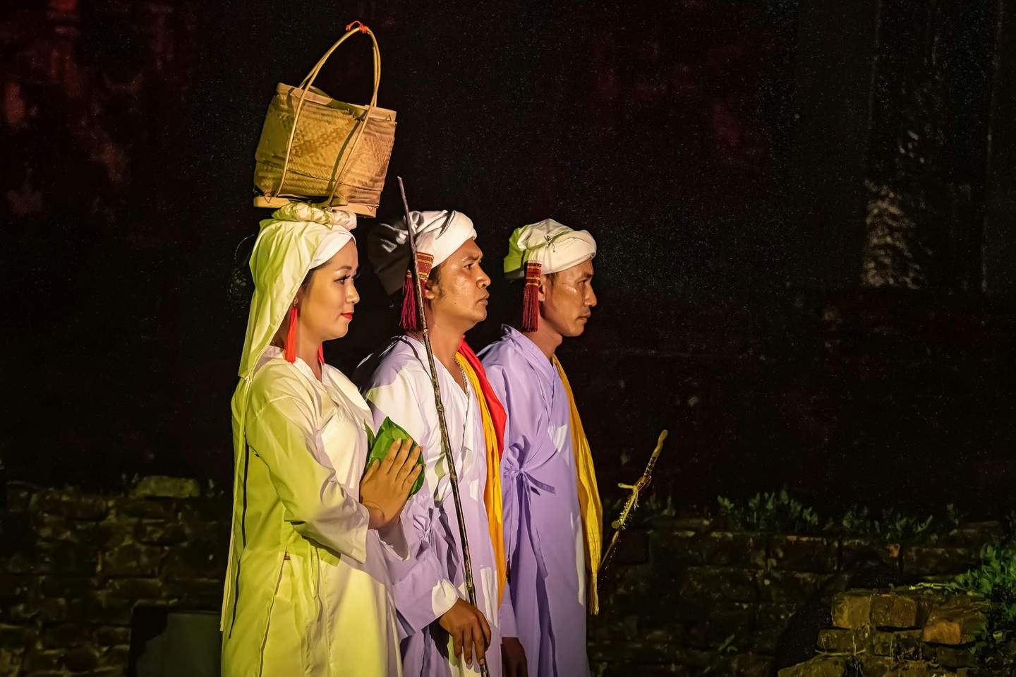 Three people in traditional attire, one with a basket on their head, standing in profile in a dimly lit setting.