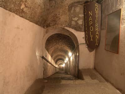 Naples Underground: Skip The Line Ticket + Guided Tour A long, narrow stone corridor with arched ceilings and wall lamps leads downwards; signs read "Napoli Sotterranea."