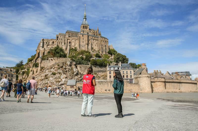 Mont Saint-Michel: Toegang + Vervoer vanuit Parijs Twee mensen staan voor de Mont Saint-Michel onder een blauwe hemel. De een draagt een rood vest en op de achtergrond zijn toeristen te zien.