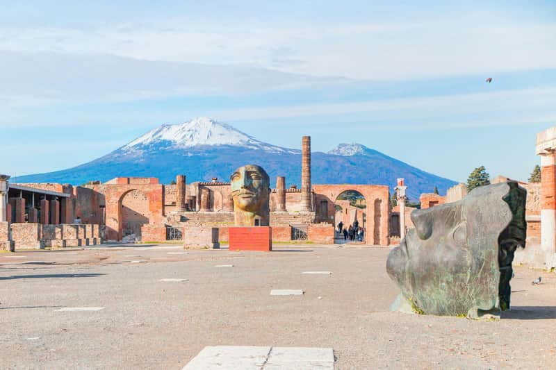 Pompeii: Skip The Line Ticket + Guided Tour Ancient ruins with large sculpted heads and Mount Vesuvius in the background under a clear sky.