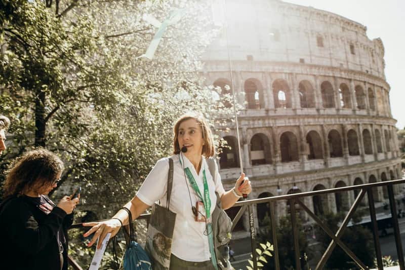 Colosseum, Arena, Roman Forum & Palatine Hill: Guided Tour A woman wearing a microphone headset leads a tour near an ancient structure, with trees and sunlight in the background.