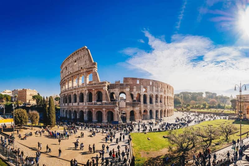 Colosseum, Roman Forum & Palatine Hill: Entry + AI Video Guide App + Map The Colosseum in Rome under a clear blue sky, with many tourists walking around the surrounding area.