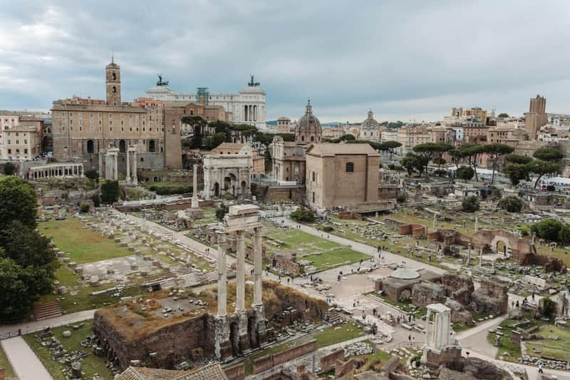 Rome: Guided Tour + Colosseum, Vatican Museums & Sistine Chapel Visit Ruins of ancient Roman buildings and columns in an open area, with modern city structures in the background under a cloudy sky.