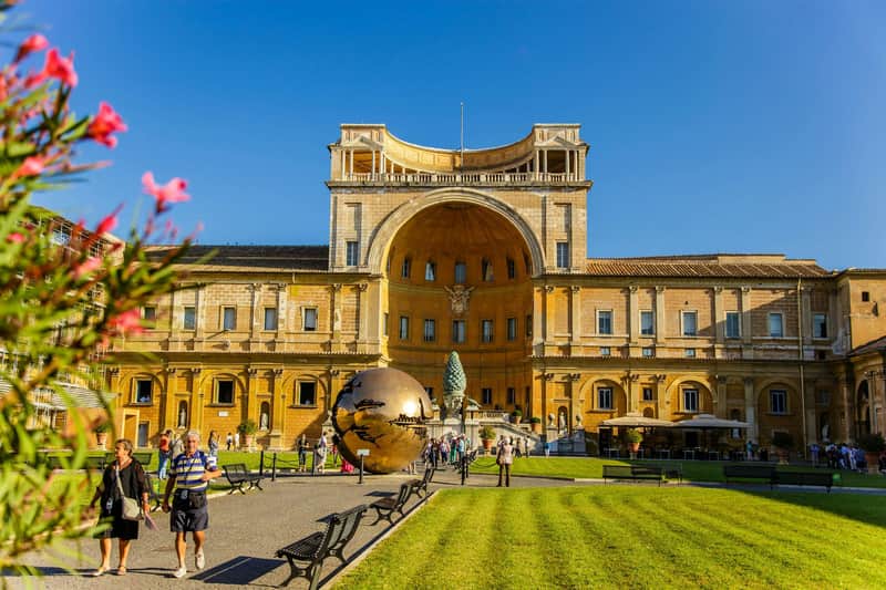 Vatican Museums, Sistine Chapel & Rome Pantheon: Fast Track Ticket People walking in a courtyard with a large bronze sphere sculpture, surrounded by historic buildings and green lawns; blue sky overhead.