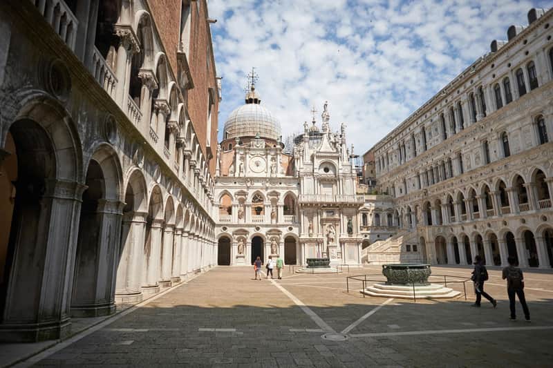 Doge's Palace: Fast Track Ticket Three people walk in a large, ornate courtyard with domed and arched architecture under a partly cloudy sky.