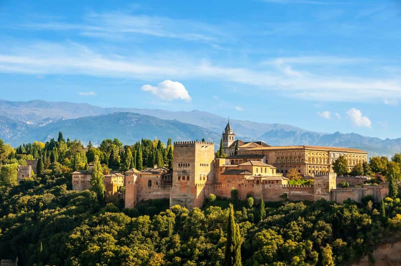 Alhambra & Nasrid Palaces: Skip The Line Ticket + Guided Tour A historic fortress with stone towers and buildings is surrounded by lush greenery, with mountains in the background under a blue sky.