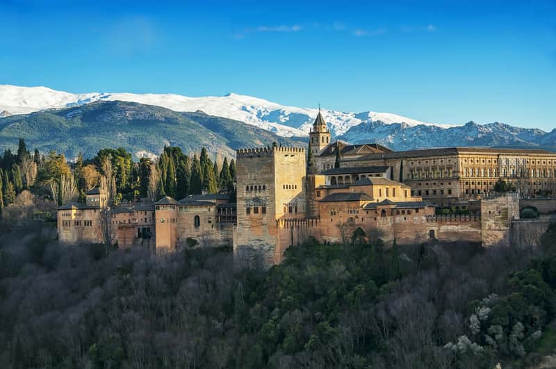 Alhambra & Nasrid Palaces: Skip The Line Ticket + Small Group Guided Tour A historic fortress with tower and buildings on a wooded hill, set against a backdrop of snow-capped mountains and a clear blue sky.