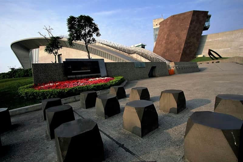 Shihsanhang Museum of Archaeology: Entry Ticket A modern architectural scene featuring angular brown structures, a garden with flowers, and geometric stone seating.