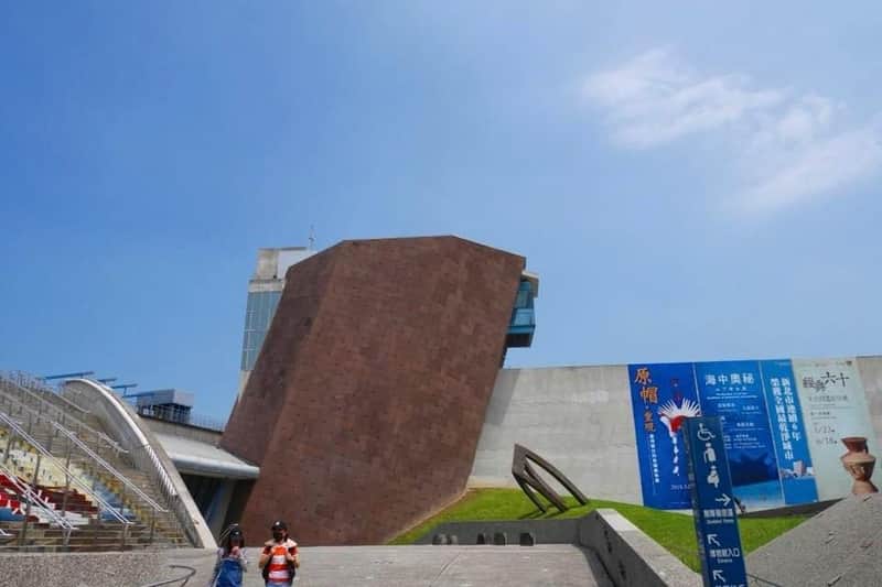 这栋现代建筑结构呈棕色棱角，白色墙壁相邻，位于晴朗的蓝天下。 两个人走在前台。