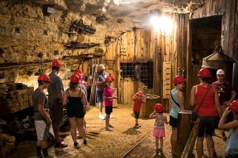 Visita guiada a la mina y viaje en tren Personas con cascos rojos exploran una cueva subterránea con paredes de madera y equipos de minería.