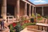 A courtyard with tall, stone columns, vibrant red flowers, and green shrubs. A few people stand in the shaded area.
