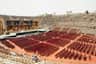 A large, empty amphitheater with rows of red chairs facing an elaborate stage. Few people are scattered in the seating area.