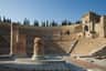 Museum of the Roman Theatre of Cartagena: Entry Ticket Ancient amphitheater with stone steps, columns in the foreground, and cypress trees on the horizon. Clear blue sky.