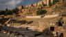Málaga Roman Theatre & Alcazaba: Guided Tour Ancient stone amphitheater with tiered seating, surrounded by ruins and vegetation under a partly cloudy sky.