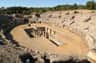 Ancient amphitheater ruins surrounded by rocky terrain and trees, with a partially deteriorated central structure.